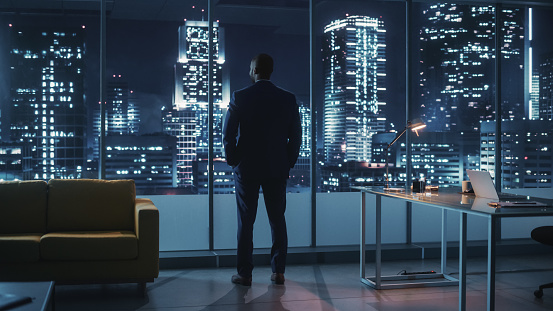 Night Office: Successful Black Businessman Wearing Suit Standing, Looking out of the Window on a Big City. African-American CEO Thinking of Environmental, Social, Corporate Governance for e-Business