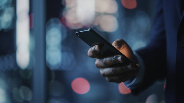 istock Night Office: Successful Black Businessman Wearing Suit Standing, Using Smartphone. CEO Browsing Internet, Managing Social Media Strategy in e-Commerce Software. Focus on Hand with Mobile Phone 1357661248