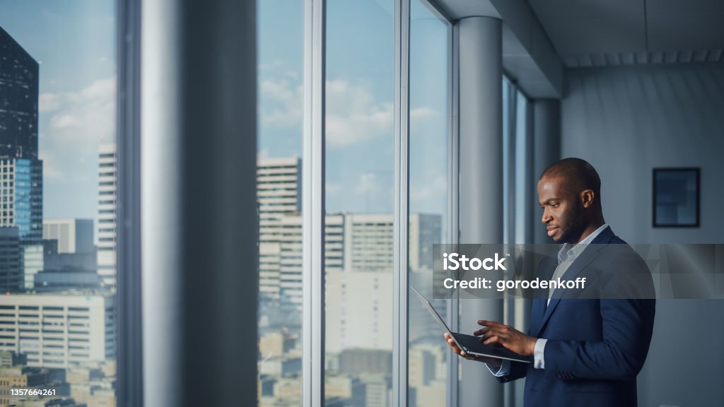 Thoughtful Black Businessman in a Tailored Suit Using Laptop while Standing in Office Near Window on Big City. Successful Corporate Top Manager Doing Data Analysis for e-Commerce Startup Business Stock Photo