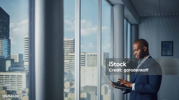 Thoughtful Black Businessman In A Tailored Suit Using Laptop While Standing In Office Near Window On Big City Successful Corporate Top Manager Doing Data Analysis For Ecommerce Startup Stock Photo - Download Image Now