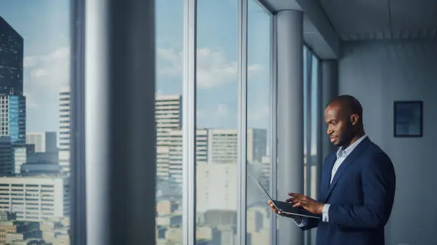 Photo of Thoughtful Black Businessman in a Tailored Suit Using Laptop while Standing in Office Near Window on Big City. Successful Corporate Top Manager Doing Data Analysis for e-Commerce Startup