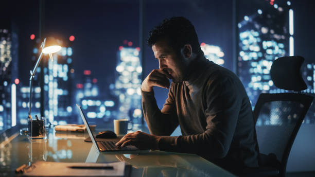 ritratto di un uomo d'affari di successo riflessivo che lavora sul computer portatile nel suo ufficio della grande città di notte. energetic digital entrepreneur fa analisi dei dati per la strategia di e-commerce. - lavoro straordinario foto e immagini stock