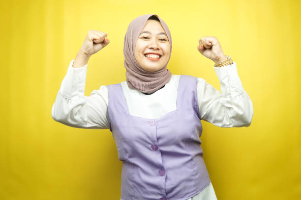 Beautiful young asian muslim woman smiling confident, enthusiastic and cheerful with  hands clenched, sign of success, punching, fighting, not afraid, isolated on yellow background stock photo