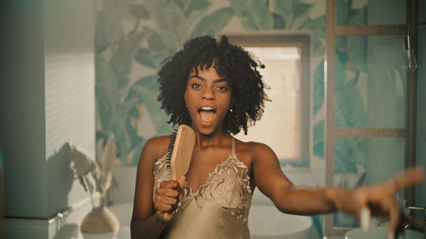 멋진 느낌. 아프리카 민족 여성은 아침 욕실 루틴을 즐기고 거울 앞에서 재미를. 헤어 브러시에 노래하고 적극적으로 춤을 - african ethnicity beauty curly hair confidence 뉴스 사진 이미지