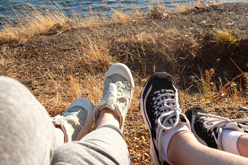 Two pairs of female legs in sports sneakers on the seashore. Female feet seen from above. Girls sitting on a hill overlooking the ocean