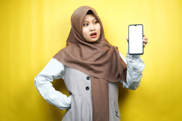 Beautiful young asian muslim woman shocked, surprised, wow expression, hand holding smartphone with white/blank screen, promoting app, promoting product, presenting something, isolated on yellow background stock photo
