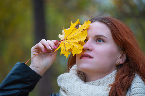 portrait of a red - haired smiling girl in a jacket and scarf with maple leaves in her hand . against the background of autumn nature, the concept of human emotion.