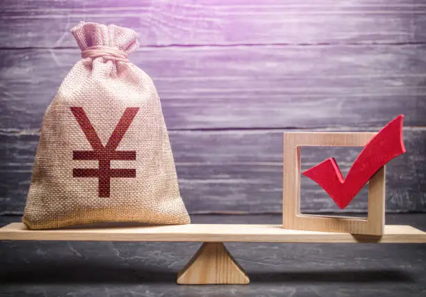 Photo of Red vote tick and a chinese yuan or japanese yen money bag on scales. Concept of lobbying for decisions and laws. Estimating cost of making a decision and consequences in the future. Corruption risks.