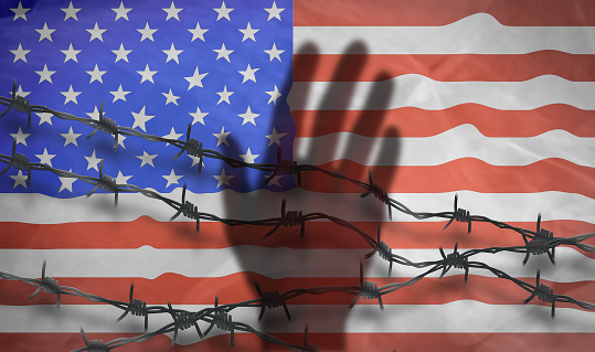 The shadow of the hand behind the barbed wire on the background of the American flag.Metal fence with barbed wire on a USA flag. Separation concept, borders protection.