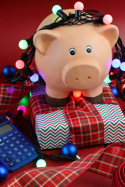 close-up image of piggy bank wrapped round with illuminated string fairy lights on wrapped christmas gift with tartan ribbon bow, blue calculator, red background, focus on foreground, christmas budget, bills, debts and finances concept - bringing home the bacon image high angle view vertical imagens e fotografias de stock