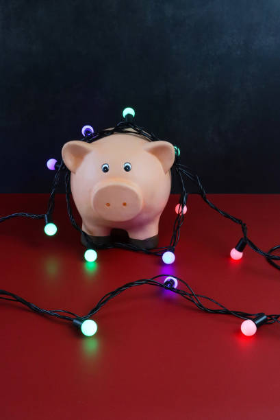 image of piggy bank wrapped round with illuminated string fairy lights on red surface, focus on foreground, black background, christmas budget, bills, debts and finances concept - bringing home the bacon image high angle view vertical imagens e fotografias de stock