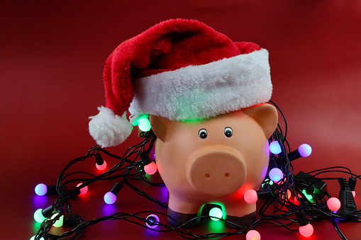 Stock photo showing a close-up view of ceramic piggy bank surrounded with illuminated Christmas string fairy lights against a red background. This is a concept picture designed to suggest Christmas finances and the general seasonal debt.