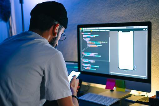 Young man mobile developer writes program code on a computer and check app in smartphone, programmer work in home office.