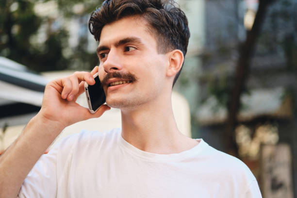 istock Portrait of young handsome mustache man in withe T-shirt talking on cellphone dreamily looking aside on city street 1357697504