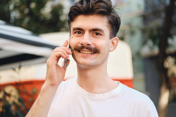 istock Portrait of young smiling mustache man in withe T-shirt talking on cellphone happily looking in camera on city street 1357697580