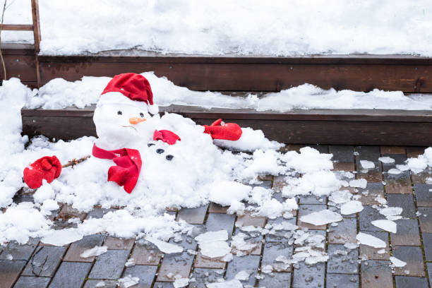Unhappy snowman in knitted mittens, red scarf and cap is melting  outdoors, snowy background stock photo