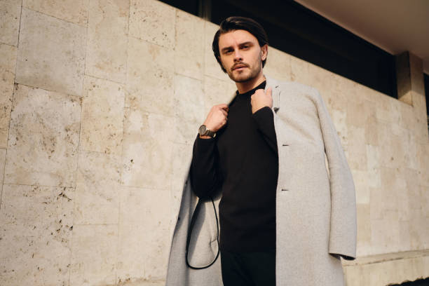 Young serious stylish brunette man with coat on shoulders thoughtfully looking in camera outdoor Young serious stylish brunette man with coat on shoulders thoughtfully looking in camera outdoor men fashion model stubble serious stock pictures, royalty-free photos & images