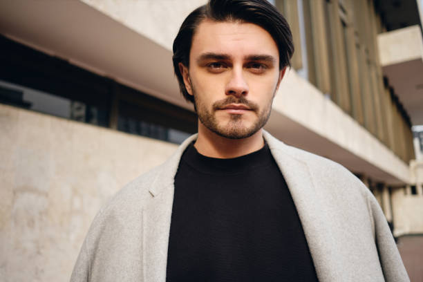 istock Young handsome stylish man with coat on shoulders thoughtfully looking in camera outdoor 1357708122