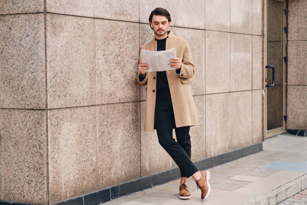 istock Young handsome confident man in beige coat thoughtfully reading newspaper outdoor 1357711290
