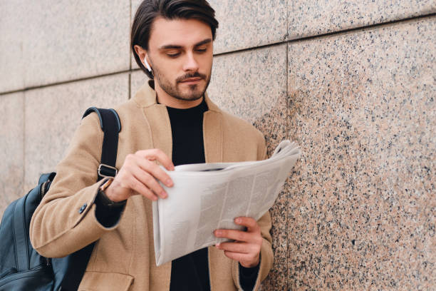istock Young confident stylish man in beige coat with backpack reading newspaper outdoor 1357712408