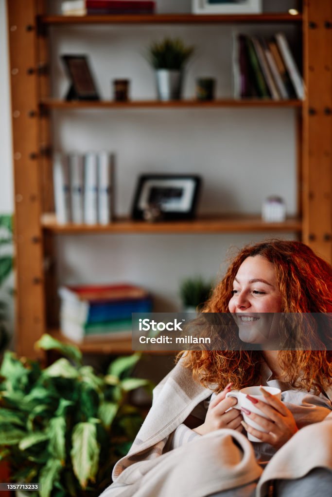 A redhead drinking coffee at home in the morning. A happy ginger girl sitting at home, holding a nice warm cup of morning coffee and enjoying free time. Domestic Life Stock Photo