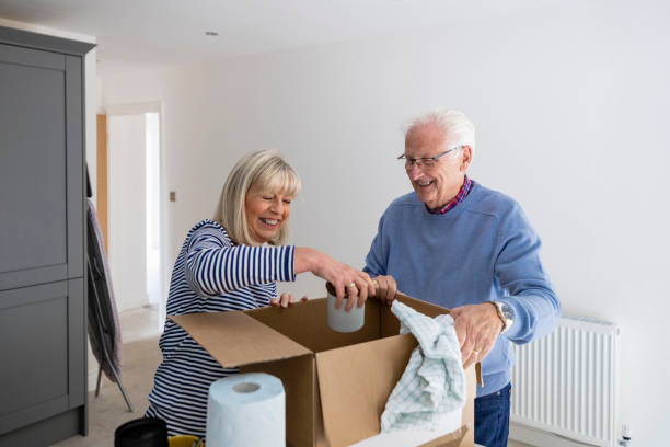 Moving House is Fun A senior couple unpacking a cardboard box filled with their kitchen belongings in the house they have just bought together. The woman is holding a mug while her husband is smiling and watching her. seniors-moving-house stock pictures, royalty-free photos & images