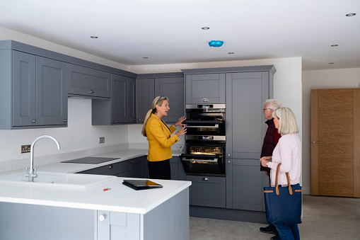 A real estate agent showing a senior couple around a new home in Northumberland, they are considering buying it. She is showing them the kitchen and talking to them about the ovens.
