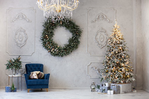 Modern stylish Christmas interior with christmas tree and gifts, blue armchair, huge wreath on the wall. Christmas decor.