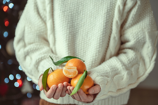 Child hands in knitted sweater holding fresh mandarins with green leaves with bokeh on background