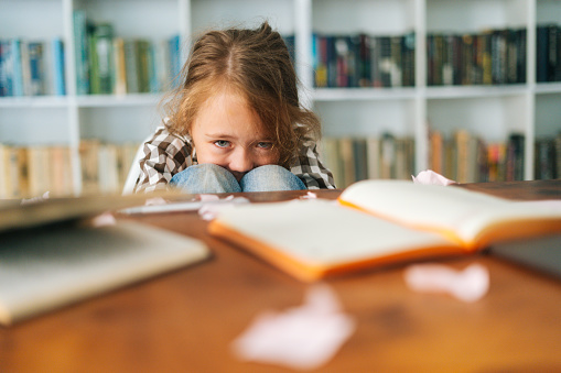 Medium shot portrait of tearing primary child school girl sitting alone hugging knees in front of desk with difficult homework, looking at camera, blurred background, selective focus.