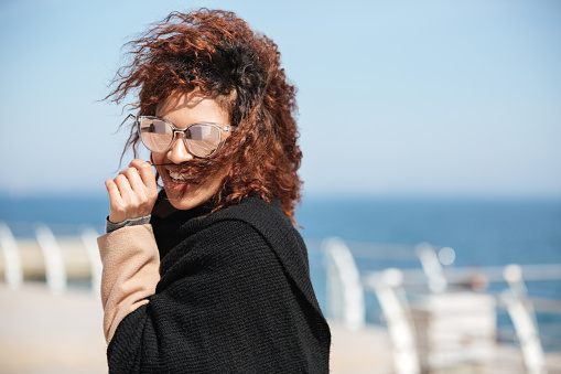 African woman in glasses laughing and holding hairs while walking near sea