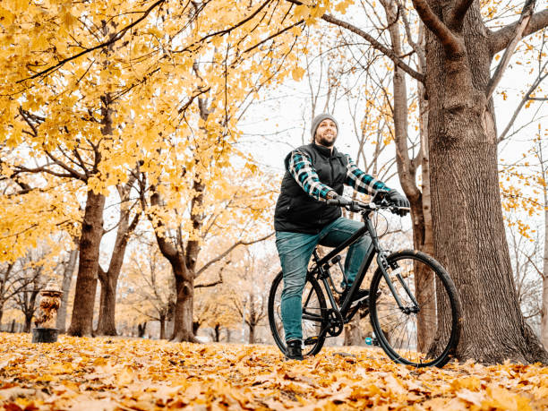 Young man amputee with prosthetic arm riding a bike in public park. Dressed in casual clothes with plaid shirt, vest and jeans. Exterior of city park in the fall with season colours of the leafs.