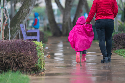 Photo of 3,5 years old girl holding her mother's hand and walking in public park. She is wearing a hooded pink raincoat and rubber boots. Shot in outdoor with a full frame mirrorless camera.