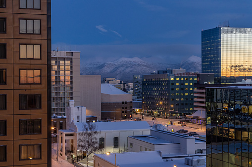 View of Downtown Anchorage on a cold winter evening. The orange of sunset is reflected in the glass of office towers. Snow covered mountains are in the distance.