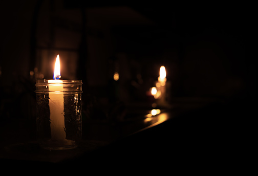 2 candles are inside a mason jar. Pitch black room except the candle lights. Selective focus on first candle with bokeh lights in the back.