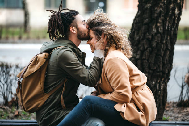 A young man kissing his girlfriend on the forehead while they are sitting on a bench at the park.