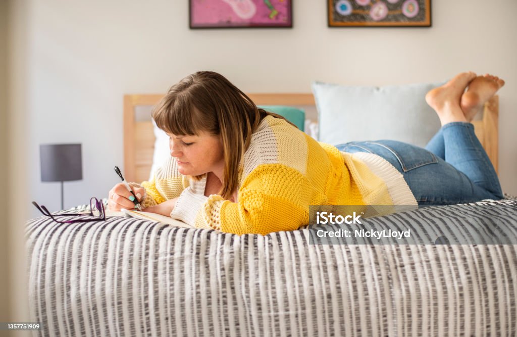 Woman lying on he bed and writing in a journal Woman lying down on her bed at home at the weekend and writing down her thoughts in a journal Copy Space Stock Photo