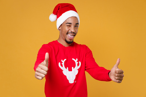 Blinking young Santa african american man 20s in red sweater with deer Christmas hat showing thumbs up isolated on yellow background studio portrait. Happy New Year celebration merry holiday concept