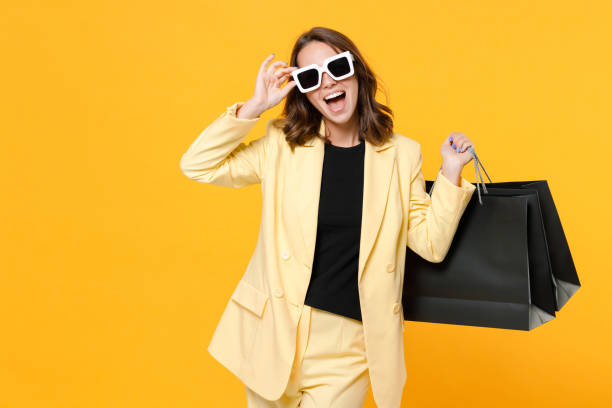 istock Cheerful young brunette woman 20s in basic light suit jacket eyeglasses hold package bags with purchases after shopping looking camera isolated on yellow background studio portrait. Black friday sale. 1357757096