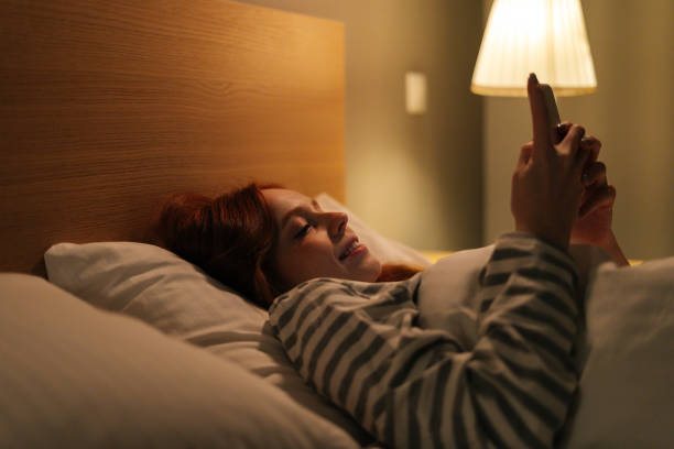 gros plan d’une jeune femme joyeuse utilisant un smartphone, regardant à l’écran, tapant un message en ligne sur les médias sociaux, allongée sur son lit tard dans la nuit. - hotel wireless technology bedroom hotel room photos et images de collection