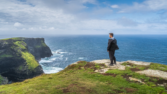 Man standing on the top of iconic Cliffs of Moher and admiring spectacular view, popular tourist attraction, Wild Atlantic Way, County Clare, Ireland