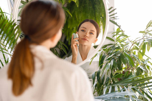 istock Young woman wearing bathrobe looking at herself in the mirror while using silicone face brush to cleanse skin, standing in bathroom decorated with green plants 1357768782