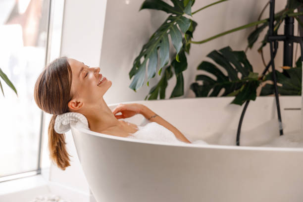 istock Side view of cheerful young woman smiling while taking a bubble bath in bathroom decorated with tropical plants 1357768843