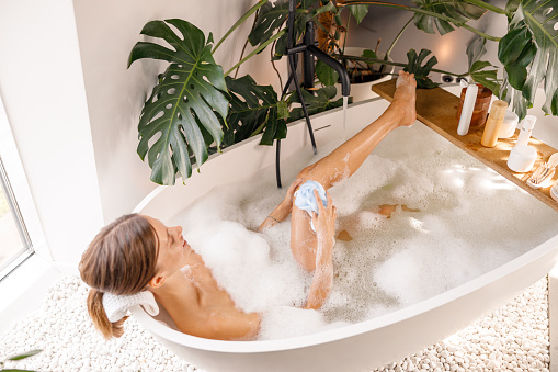 Sensual young woman using sponge, enjoying bathing with soap foam at luxury spa resort. Wellness, body care concept