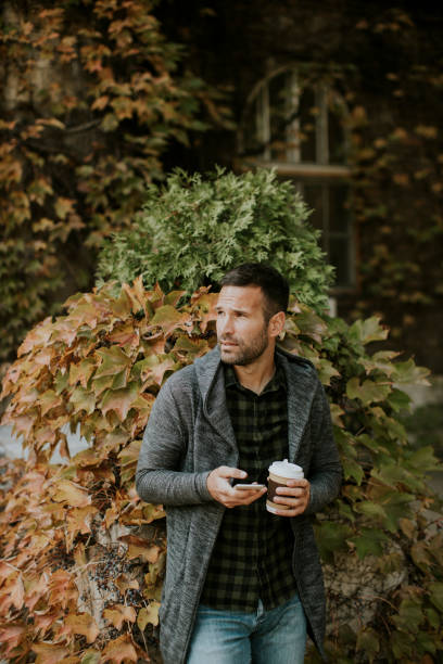 istock Young man using mobile phone and holding takeaway coffee in the park 1357775744