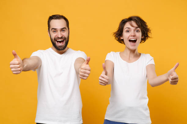 excited young couple friends bearded guy girl in white blank empty t-shirts posing isolated on yellow orange wall background in studio. people lifestyle concept. mock up copy space. showing thumbs up. - made man object imagens e fotografias de stock