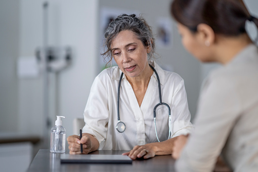 A mature female doctor points to the screen of her tablet laying out on the table in front of her as she shares her patients test results with her.  She is wearing a white lab coat and has a stethoscope around her neck as she talks with the female patient of Asian decent.
