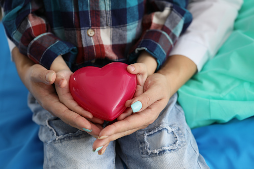 Close-up of woman and child holding red heart on palms symbol of love. Parent and son share message of charity and donation. Good deed, save life concept