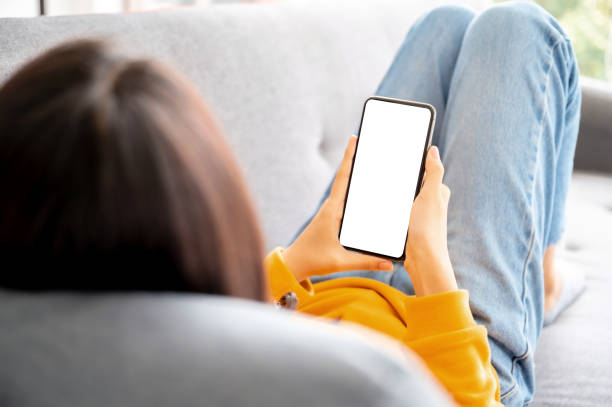 istock Mock up image white screen mobile smartphone. Woman using smart phone and relaxing on sofa. Clipping path included device screen 1357798037