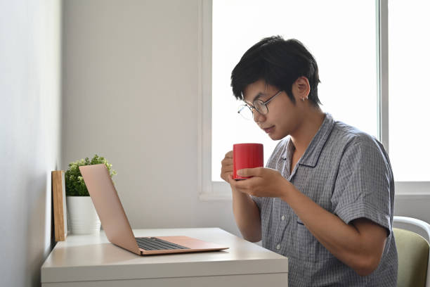 istock Peaceful asian man in pajamas using computer laptop in bedroom and drinking coffee. 1357813122
